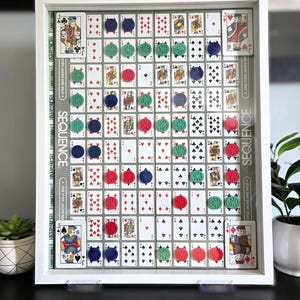 May include: A framed board game, "Sequence", with playing cards and colored game pieces. The game board is a grid of playing cards, with red, green, and blue game pieces placed on the cards. The frame is white.