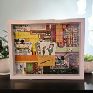 May include: A framed shadow box featuring a vintage Clue board game. The artwork includes game cards, a game board, and the text "CONFIDENTIAL". The frame is white, and the game pieces are visible.