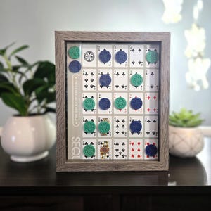 May include: A framed game board featuring playing cards with blue and green game pieces. The game board is in a wooden frame. The text "TWO EYED JACKS ARE WILD" and "SEQU" are visible on the board.