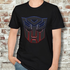 Könnte beinhalten: Schwarzes T-Shirt mit einem Autobot-Logo mit Karbonfaser-Textur in Rot und Blau. Das Logo ist ein stilisiertes Gesicht mit geometrischen Formen. Das T-Shirt hat einen Rundhalsausschnitt und ist aus weichem Material.