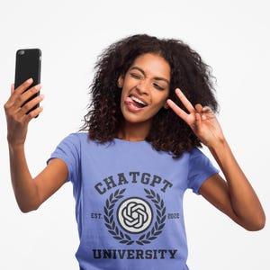 May include: A light blue t-shirt with the text "CHATGPT UNIVERSITY" in a collegiate style design. The person is holding a black smartphone and making a peace sign with their fingers.