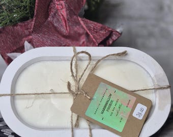 May include: A white, oval-shaped wooden tray holds a cream-colored candle, tied with twine. A brown tag reads "Marshmallow Fireside" and "16 oz." The candle sits on a black and white checkered cloth, with a red decorative element in the background.