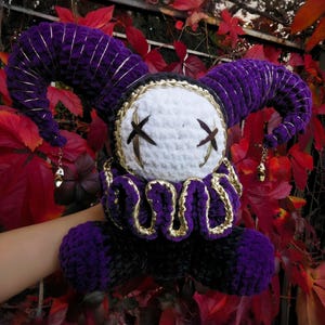 May include: A plush jester doll crafted from purple and black yarn, featuring a white round face with stitched dark brown X-shaped eyes. The doll has two large curved horns with gold thread accents, a ruffled collar, and small gold bells at the tips.