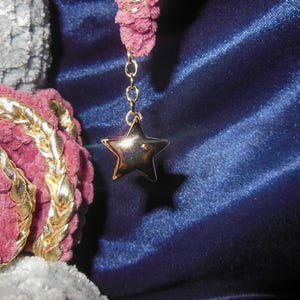 May include: A gold-toned star charm suspended from a delicate chain attached to a textured, mauve-colored yarn element. The star features a polished, reflective surface, contrasting with the soft, looped appearance of the surrounding yarn.