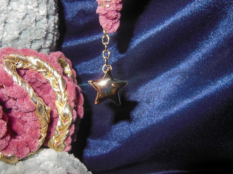 May include: A gold-toned star charm suspended from a delicate chain attached to a textured, mauve-colored yarn element. The star features a polished, reflective surface, contrasting with the soft, looped appearance of the surrounding yarn.