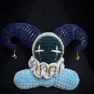 May include: A plush toy featuring a dark green round head with white stitched eyes, topped with two large dark blue curved horns accented with gold thread. It has a white ruffled collar with gold trim and a light blue base, with gold star charms.