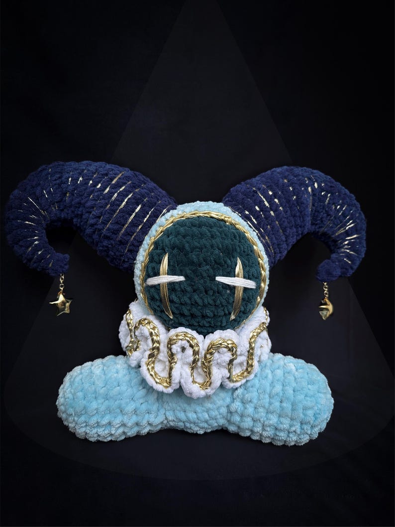 May include: A plush toy featuring a dark green round head with white stitched eyes, topped with two large dark blue curved horns accented with gold thread. It has a white ruffled collar with gold trim and a light blue base, with gold star charms.