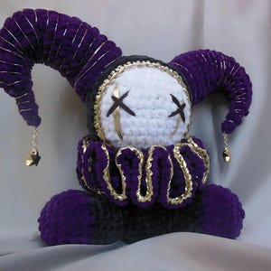 May include: A plush jester doll crafted from purple and black yarn, featuring a white face with black stitched X-shaped eyes. The doll wears a ruffled collar with gold trim and has two curved horns topped with small gold star charms.