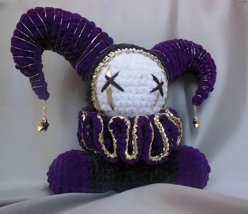 May include: A plush jester doll crafted from purple and black yarn, featuring a white face with black stitched X-shaped eyes. The doll wears a ruffled collar with gold trim and has two curved horns topped with small gold star charms.