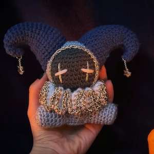 May include: A handmade crochet doll in the shape of a jester. The doll features a dark blue hat with dangling star charms, a gray face with cross-shaped accents, and a gold chain collar. The doll is held in a hand against a dark background.
