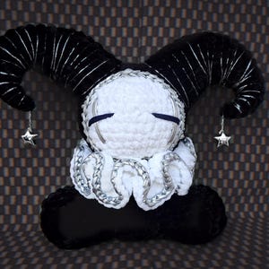 May include: A plush doll featuring a round white crocheted head with closed eyes, large black curved horns with silver accents, and a white ruffled collar trimmed in silver. Two small silver star charms hang from the tips of the horns.