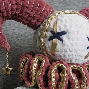 May include: A plush doll featuring a white round head with dark blue stitched X-shaped eyes, accented by gold trim. It has two large, curved, dusty rose plush horns wrapped in gold wire, each ending in a small gold star charm. A ruffled collar in dusty rose and gold surrounds the neck.