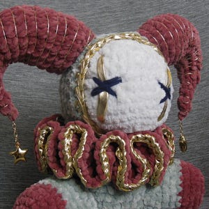 May include: A plush doll featuring a white face with dark blue stitched X-shaped eyes and gold accents. It has two large, curved, dusty rose horns wrapped in gold thread, a ruffled collar with gold trim, and gold star charms hanging from the horns.