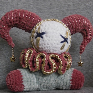 May include: A plush jester doll crafted from soft yarn, featuring a white round head with dark blue stitched X-shaped eyes, two curved maroon horns with gold trim, a ruffled collar, and a light gray body with maroon accents and gold star charms.