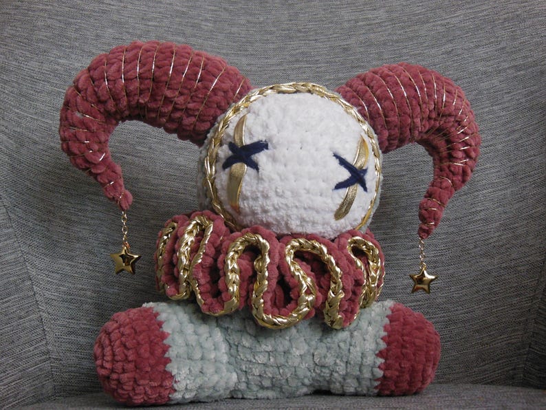 May include: A plush jester doll crafted from soft yarn, featuring a white round head with dark blue stitched X-shaped eyes, two curved maroon horns with gold trim, a ruffled collar, and a light gray body with maroon accents and gold star charms.
