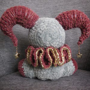 May include: A plush toy featuring a round gray body with two large, curved, dark red horns wrapped in gold thread. A ruffled dark red collar with gold trim encircles the base. Two small gold star charms hang from the tips of the horns.
