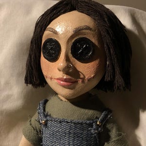 May include: A handmade doll with button eyes and stitched mouth. The doll has dark hair and is wearing a denim overall top over a green shirt. The doll's face is painted with a pale complexion and rosy cheeks.