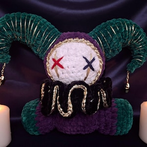 May include: A plush jester doll featuring a white round head with a red stitched X eye and a dark blue stitched X eye. The doll has dark green velvet horns with gold trim, a black ruffled collar with gold accents, and a purple and green body.