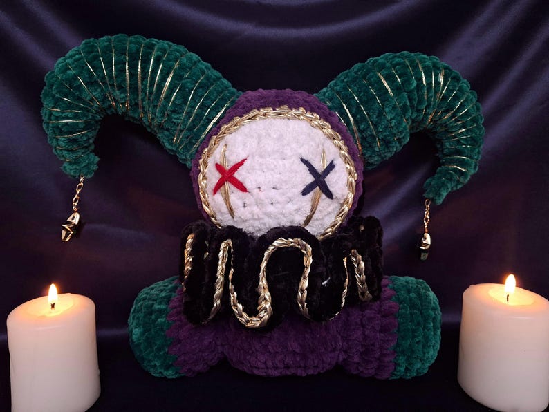 May include: A plush jester doll featuring a white round head with a red stitched X eye and a dark blue stitched X eye. The doll has dark green velvet horns with gold trim, a black ruffled collar with gold accents, and a purple and green body.