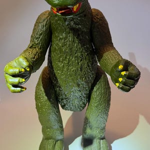 Puede incluir: Un juguete de monstruo verde grande con una superficie texturizada. El juguete tiene garras y dientes amarillos y un collar rojo. El monstruo tiene una expresión amenazante y está en posición de pie. El juguete es probablemente un objeto de colección o vintage.