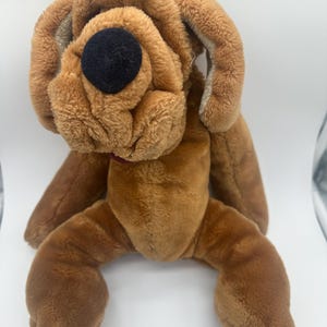 May include: A plush brown dog toy with floppy ears and a large black eye. The stuffed animal has a soft, textured appearance and a small red collar. The dog is sitting with its legs extended.