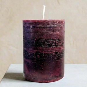 May include: A cylindrical pillar candle in shades of deep burgundy and purple. The candle has a textured surface with a visible wick at the top. The candle is approximately 4 inches tall.