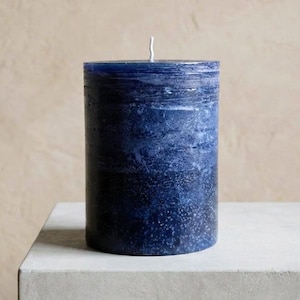 May include: A cylindrical, dark blue candle with a textured surface. The candle has a white wick and sits on a light gray surface. The color transitions from a deep indigo at the base to a lighter shade at the top.