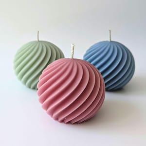 May include: Three spherical candles in shades of green, pink, and blue. Each candle has a spiral design and a white wick. The candles are arranged on a white surface.