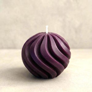 May include: A spherical candle in a deep purple color featuring a distinct spiral or twisted ribbed texture across its entire surface. A single white cotton wick protrudes from the center top of the rounded wax form, which has a smooth finish.