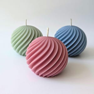 May include: Three spherical candles in shades of green, pink, and blue. Each candle has a spiral design and a single wick. The candles are arranged on a white surface, creating a simple and elegant aesthetic.