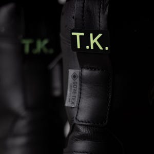 Könnte beinhalten: Nahaufnahme von schwarzen Lederstiefeln mit den Buchstaben "T.K." in Neongrün auf der Ferse. Die Stiefel haben ein "GORE-TEX"-Etikett. Das Bild ist dunkel und konzentriert sich auf die Details der Stiefel.