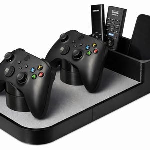May include: Black gaming controller organizer with two controllers, two remotes, and a smartphone. The organizer has a two-tiered design with a gray surface and is made of black material.