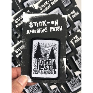 May include: A white iron-on patch with black outlines of two pine trees, a sun, and clouds. The text "GET LOST" is in black and is layered over the trees. The patch is on a black card with white text that reads "STICK-ON ADVENTURE PATCH".