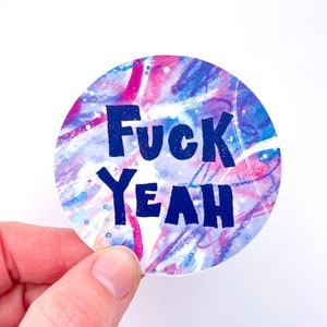 May include: A round sticker with a white background and a blue, pink, and purple watercolor design. The sticker says "FUCK YEAH" in bold, dark blue letters.