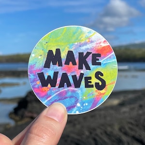 May include: A round sticker with a tie-dye background in shades of blue, green, yellow, pink, and purple. The sticker has the text "MAKE WAVES" in black.