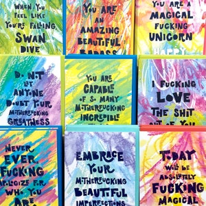 May include: A collection of colorful cards with motivational and humorous messages. The cards feature a watercolor-like background with bold black lettering. Some of the messages include "You are an amazing beautiful badass", "Never ever fucking apologize for who you are", and "Today will be absolutely fucking magical".