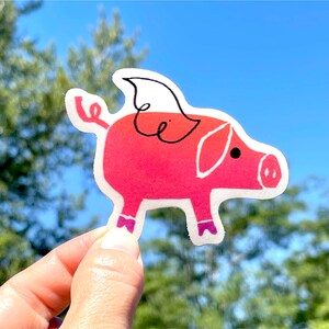 May include: A pink pig sticker with a white wing and purple shoes. The pig is holding a curled tail in its mouth.