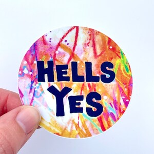 May include: A round sticker with a colorful abstract background and the text "HELLS YES" in bold, black letters.