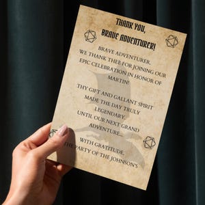 May include: A vintage-style thank you card with the text "Thank You, Brave Adventurer!" and other text. The card has a parchment-like appearance with decorative geometric shapes. A hand is holding the card.