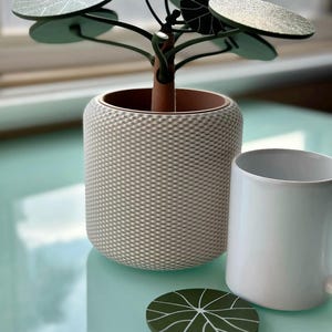 May include: A decorative plant sculpture with round green leaves featuring white vein patterns, set in a cylindrical pot with a textured cream-colored woven exterior. A plain white ceramic mug and a single matching green leaf coaster sit nearby.