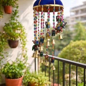 May include: A hanging wind chime with a blue velvet top and gold trim. Colorful beads and strings of small bells and elephant figurines dangle below. The wind chime is hanging on a balcony with plants.