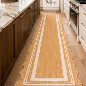 May include: A rectangular jute runner rug with a scalloped edge. The rug features a light tan color with a white border and two white stripes. The rug is placed on a hardwood floor in a kitchen.