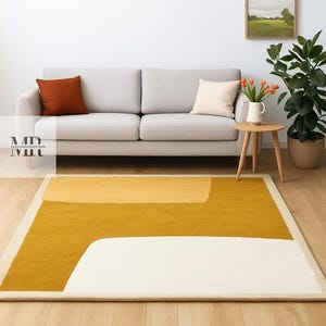 Pode incluir: Um tapete retangular com um design moderno. O tapete apresenta uma grande seção amarelo mostarda, uma seção branca e uma borda creme. Um sofá cinza com almofadas laranja e creme está ao fundo.