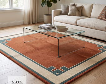 Puede incluir: Una alfombra rectangular que presenta un gran campo central de color terracota, bordeado por una banda ancha de color crema y un fino marco geométrico de color verde azulado. La alfombra muestra una textura tejida plana sobre suelo de madera.
