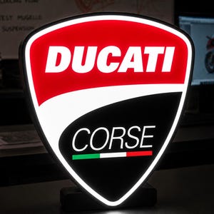 Może przedstawiać: Podświetlany szyld w kształcie tarczy z logo Ducati Corse. Górna część jest czerwona z białym napisem DUCATI, a dolna część jest czarna z białym napisem CORSE nad małym poziomym paskiem w kolorach zielonym, białym i czerwonym.