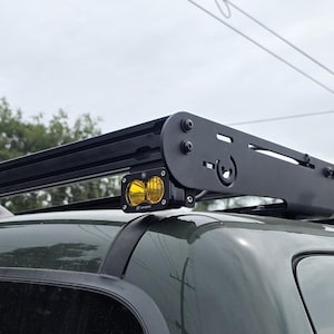 May include: Black roof rack with integrated yellow lights mounted on a dark green SUV. The rack is made of metal and has a sleek, modern design. The lights are round and provide additional illumination. The vehicle is parked outdoors under a cloudy sky.