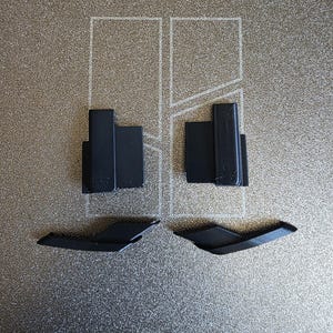 May include: Four black plastic components are arranged on a textured, light brown surface. Two rectangular pieces with a raised section are positioned at the top, and two angled, wing-shaped pieces are below.