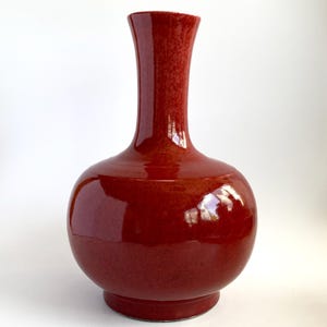 Puede incluir: Un jarrón de cerámica rojo brillante, con un cuerpo redondeado y un cuello delgado y alargado. El jarrón tiene una base ancha y una superficie lisa y reflectante, que muestra un rico tono rojo oscuro. Este jarrón decorativo es adecuado para la decoración del hogar.