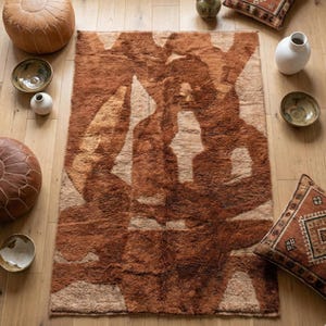 May include: A rectangular, brown and beige patterned rug with a shaggy texture. The rug features an abstract design with lighter beige shapes against a darker brown background. Decorative pillows, bowls, and poufs surround the rug.