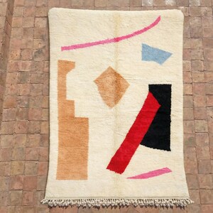 Könnte beinhalten: Cremefarbener, rechteckiger Teppich mit einem abstrakten Design. Der Teppich hat geometrische Formen in verschiedenen Farben, darunter Beige, Rot, Schwarz, Rosa und Blau. Der Teppich hat einen Fransenbesatz am unteren Rand.